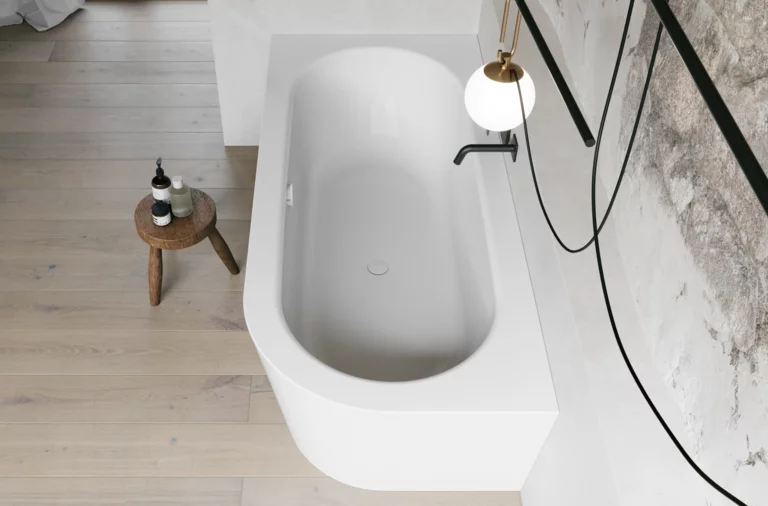 Wit inbouwbad van bovenaf in een lichte badkamer met zwarte kraan, krukje en wand in steenlook.