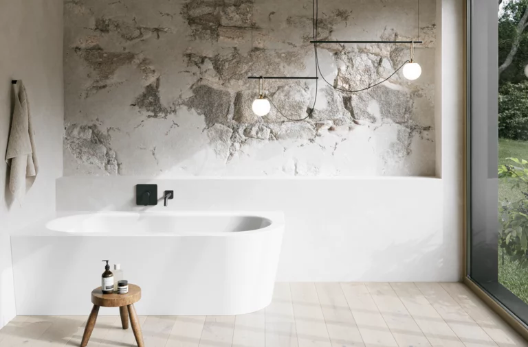 Wit inbouwbad in een lichte badkamer met wand in steenlook, krukje, handdoek en groot raam.