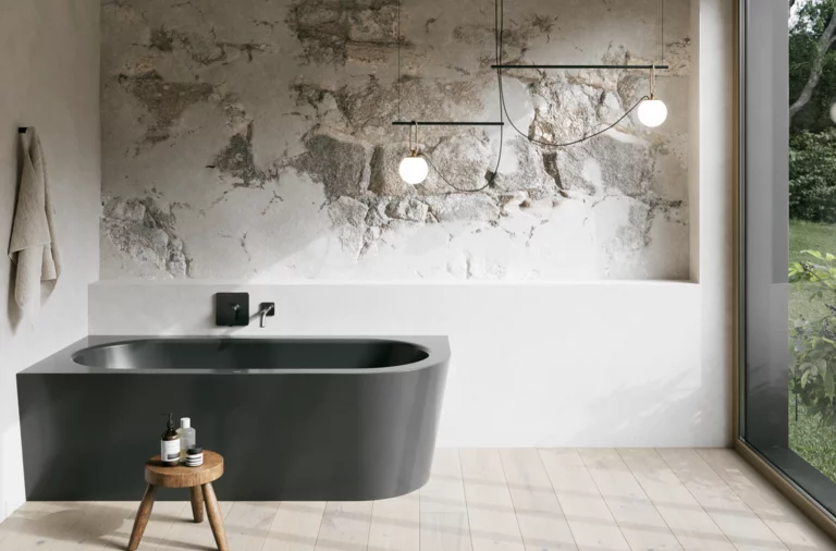 Donker inbouwbad in een lichte badkamer met wand in steenlook, krukje, handdoek en groot raam.