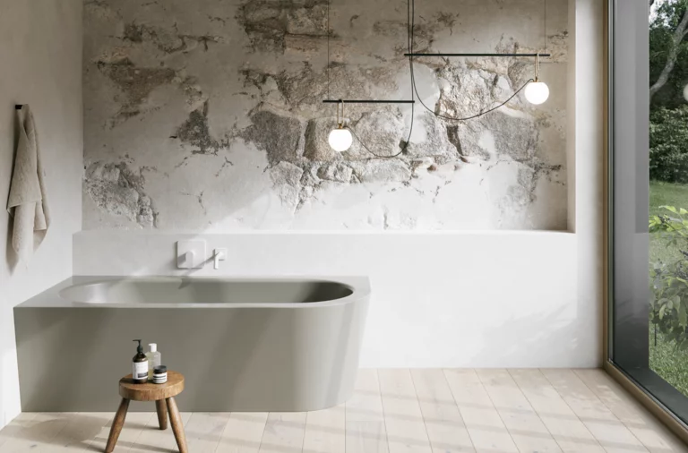 Beige inbouwbad in een lichte badkamer met wand in steenlook, krukje, handdoek en groot raam.