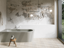 Beige inbouwbad in een lichte badkamer met wand in steenlook, krukje, handdoek en groot raam.