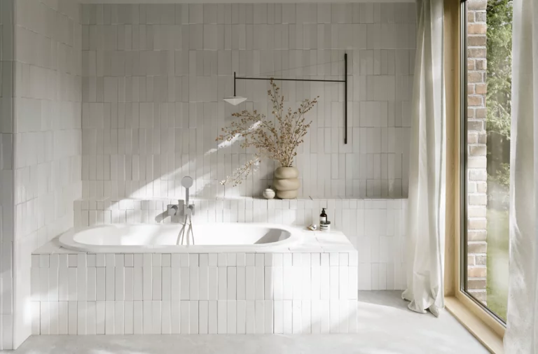 Weiße Einbau-Badewanne in hellem Bad mit Fliesenoptik, Vase, silberner Armatur und großem Fenster.