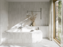 Weiße Einbau-Badewanne in hellem Bad mit Fliesenoptik, Vase, silberner Armatur und großem Fenster.
