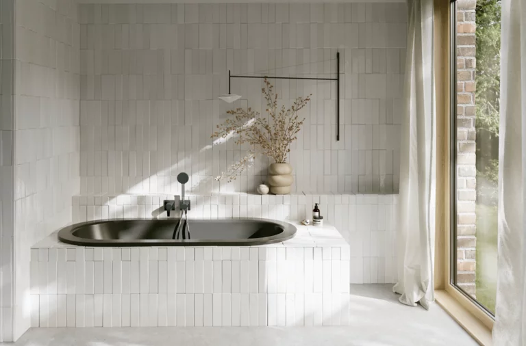 Dunkle Einbau-Badewanne in hellem Bad mit Fliesenoptik, Vase, schwarzer Armatur und großem Fenster.