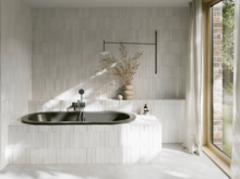 Dunkle Einbau-Badewanne in hellem Bad mit Fliesenoptik, Vase, schwarzer Armatur und großem Fenster.