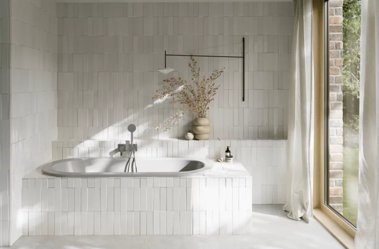 Hellgraue Einbau-Badewanne in hellem Bad mit Fliesenoptik, Vase, silberner Armatur und großem Fenster.