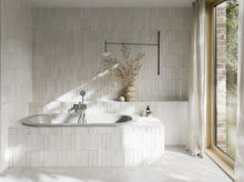 Hellgraue Einbau-Badewanne in hellem Bad mit Fliesenoptik, Vase, silberner Armatur und großem Fenster.
