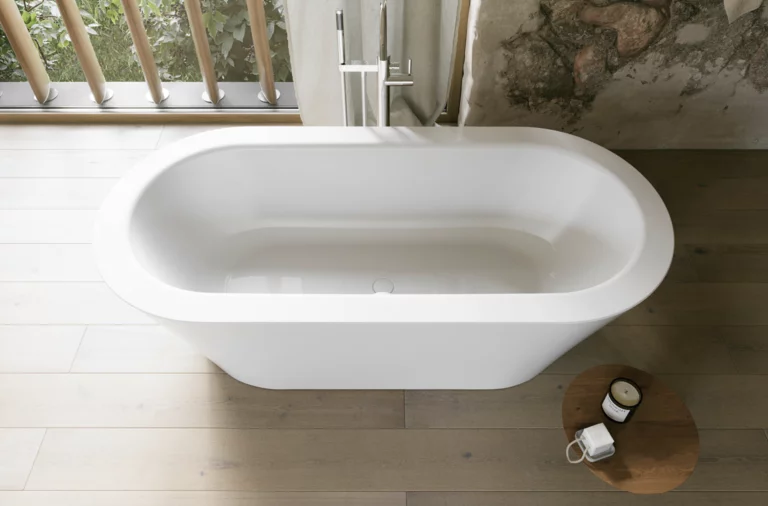 Weiße freistehende Badewanne aus Draufsicht in hellem Bad mit Holzoptik-Boden, Beistelltisch und Fenster.