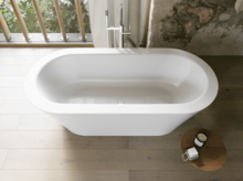 Weiße freistehende Badewanne aus Draufsicht in hellem Bad mit Holzoptik-Boden, Beistelltisch und Fenster.