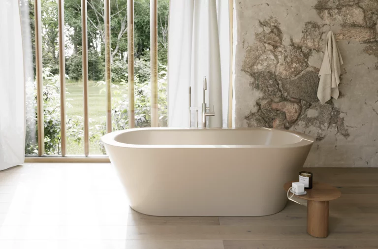Beige freistehende Badewanne in hellem Bad mit Natursteinoptik-Wand, Beistelltisch und großem Fenster.