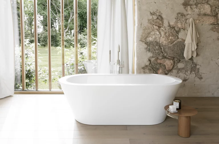 Weiße freistehende Badewanne in hellem Bad mit Natursteinoptik-Wand, Beistelltisch und großem Fenster.