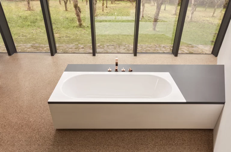 Vasca da bagno rettangolare in acciaio smaltato, parzialmente integrata in una mensola laterale, con rubinetteria, davanti a grandi finestre.