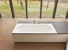 Rechteckige Stahl-Email-Badewanne, teilweise in eine seitliche Ablagefläche integriert, mit Armaturen, vor großen Fenstern.