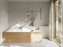 Weiße Einbau-Badewanne in hellem Bad mit heller Verkleidung, Fliesenoptik, Vase und großem Fenster.