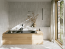 Dunkle Einbau-Badewanne in hellem Bad mit heller Verkleidung, Fliesenoptik, Vase und großem Fenster.