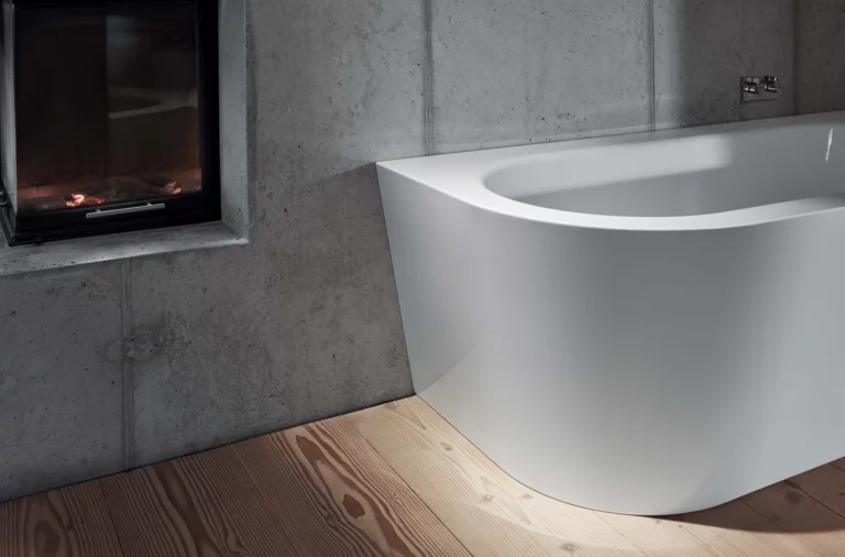 Freistehende Badewanne aus Stahl-Email mit abgerundeter Ecke, vor Betonwand und Kamin, auf Holzfußboden.