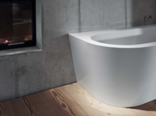 Freistehende Badewanne aus Stahl-Email mit abgerundeter Ecke, vor Betonwand und Kamin, auf Holzfußboden.