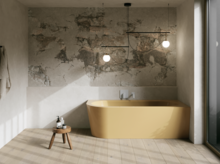 Gelbe freistehende Badewanne in hellem Bad mit Natursteinoptik-Wand, Hocker, Handtuch und großem Fenster.