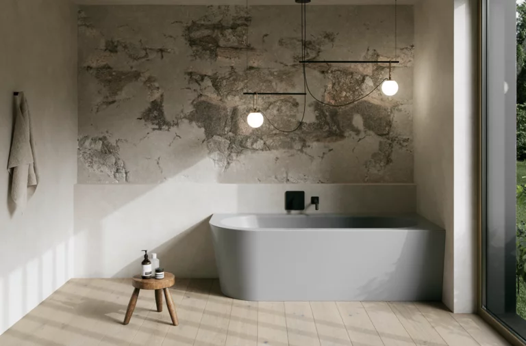 Graue freistehende Badewanne in hellem Bad mit Natursteinoptik-Wand, Hocker, Handtuch und großem Fenster.