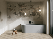 Graue freistehende Badewanne in hellem Bad mit Natursteinoptik-Wand, Hocker, Handtuch und großem Fenster.