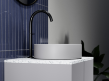 Rundes Waschbecken aus Stahl-Email auf Einbauplatte mit schwarzem Wasserhahn, blauer Fliesenwand und Pflanze im Hintergrund.