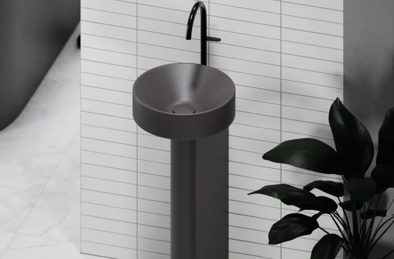 Rundes Standwaschbecken mit schwarzem Wasserhahn vor gefliester Wand, daneben grüne Pflanze.