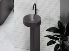 Rundes Standwaschbecken mit schwarzem Wasserhahn vor gefliester Wand, daneben grüne Pflanze.