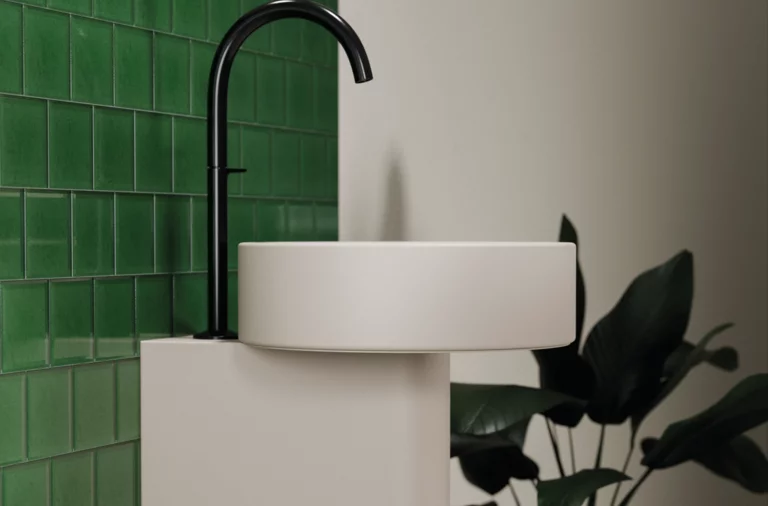 Rundes Waschbecken auf Einbauplatte mit schwarzer Armatur vor grünen Wandfliesen und Pflanze im Hintergrund.
