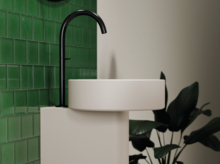 Rundes Waschbecken auf Einbauplatte mit schwarzer Armatur vor grünen Wandfliesen und Pflanze im Hintergrund.