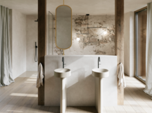 Lichte badkamer met twee beige vrijstaande wastafels, zwarte wandkranen en een grote spiegel voor een stenen wand.