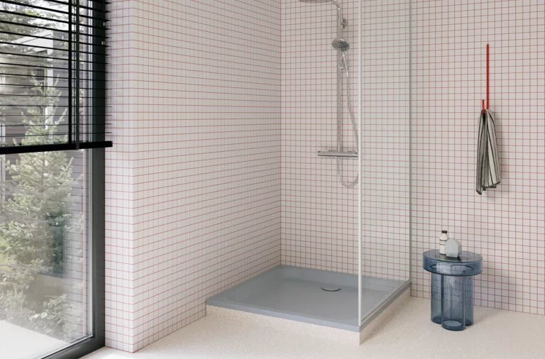 Hellgraue Duschwanne in einer modernen Dusche mit weißen Mosaikfliesen, Glasabtrennung, silberner Armatur und großem Fenster.