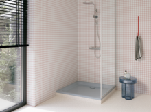 Hellgraue Duschwanne in einer modernen Dusche mit weißen Mosaikfliesen, Glasabtrennung, silberner Armatur und großem Fenster.