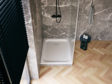 Weiße Duschwanne in moderner Dusche mit grauer Marmoroptik, Glasabtrennung, schwarzer Armatur und hellem Fischgrätboden.