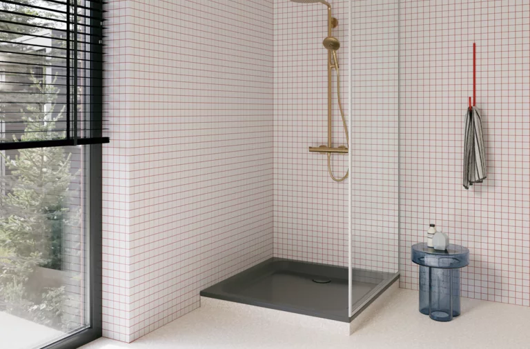 Dunkle Duschwanne in einer hellen Dusche mit weißen Mosaikfliesen, Glasabtrennung, goldfarbener Armatur und großem Fenster.