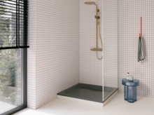 Dunkle Duschwanne in einer hellen Dusche mit weißen Mosaikfliesen, Glasabtrennung, goldfarbener Armatur und großem Fenster.