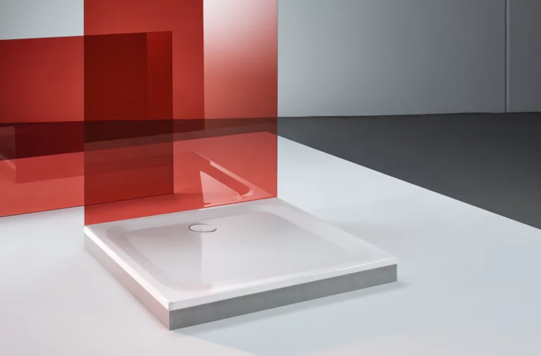 Quadratische Duschwanne aus Stahl-Email mit flachem Rand, montiert auf hellem Boden, rote Glasscheibe daneben.