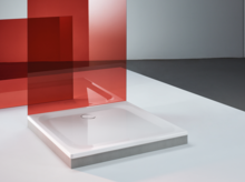 Quadratische Duschwanne aus Stahl-Email mit flachem Rand, montiert auf hellem Boden, rote Glasscheibe daneben.