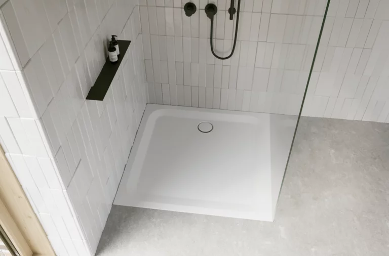 Weiße Duschwanne in moderner Dusche mit weißen Fliesen, Glasabtrennung, schwarzer Armatur und grauem Boden.