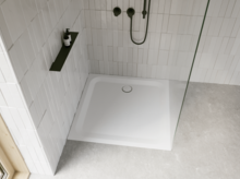 Weiße Duschwanne in moderner Dusche mit weißen Fliesen, Glasabtrennung, schwarzer Armatur und grauem Boden.