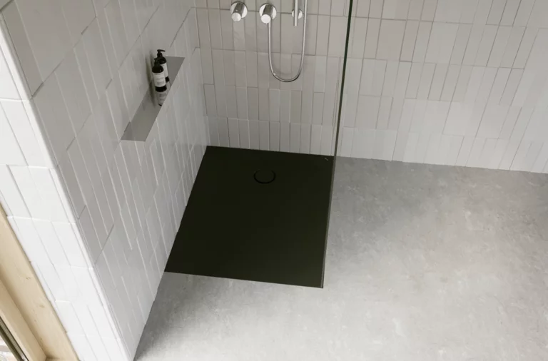 Dunkle, bodengleiche Duschwanne in moderner Dusche mit weißen Fliesen, Glasabtrennung, silberner Armatur und grauem Boden.