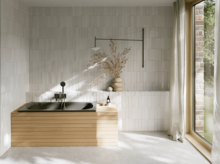 Helles Bad mit dunkel eingelassener Badewanne, heller Front, Fliesenwand, bodentiefem Fenster und Vorhang.