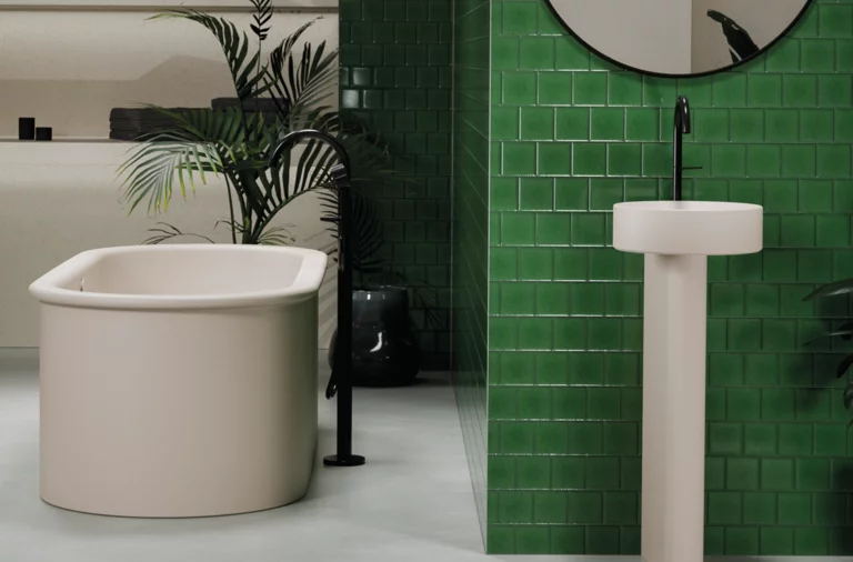 Freestanding oval bathtub made of steel enamel and round pedestal washbasin in front of green tiles.
