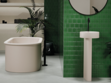 Freestanding oval bathtub made of steel enamel and round pedestal washbasin in front of green tiles.