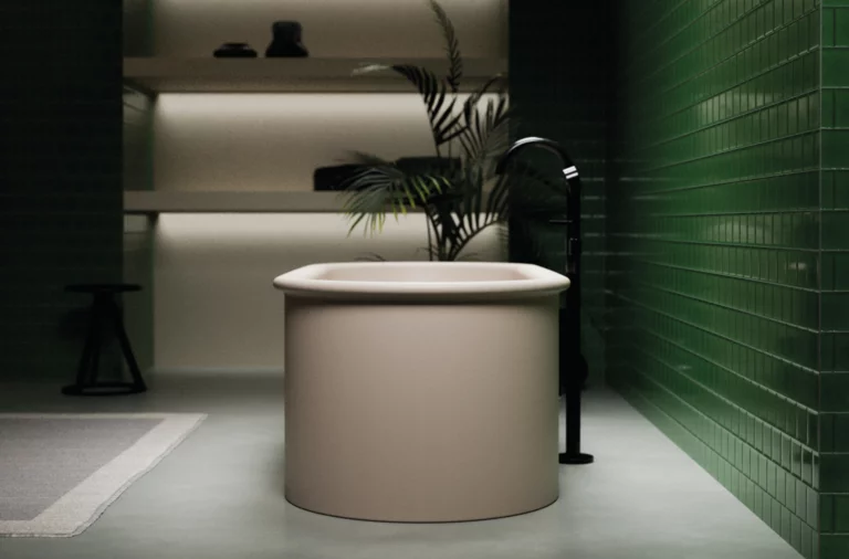 Round freestanding steel enamel bathtub in modern bathroom with green tiled wall and black floor-mounted faucet.