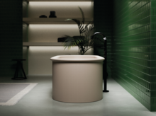 Round freestanding steel enamel bathtub in modern bathroom with green tiled wall and black floor-mounted faucet.