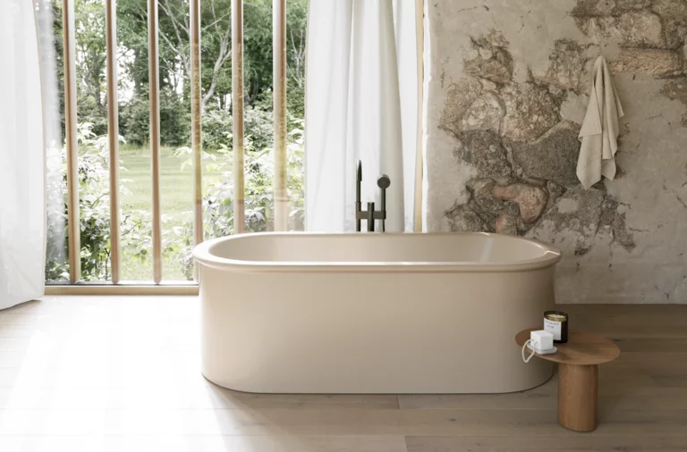 Freistehende beige Badewanne vor bodentiefen Fenstern, neben rustikaler Steinwand und kleinem Hocker.