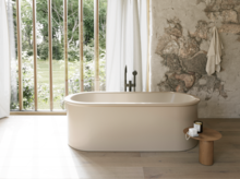 Freistehende beige Badewanne vor bodentiefen Fenstern, neben rustikaler Steinwand und kleinem Hocker.